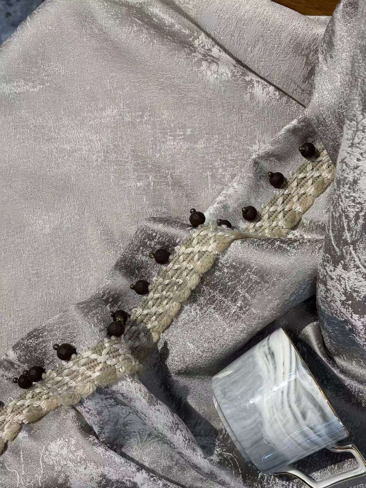 这是一张银灰色高档窗帘面料的特写图。面料表面呈现出细腻的银灰色光泽，带有不规则的纹理质感，显得非常有档次。边缘装饰有精致的米白色编织花边，并串有深褐色的圆珠，增